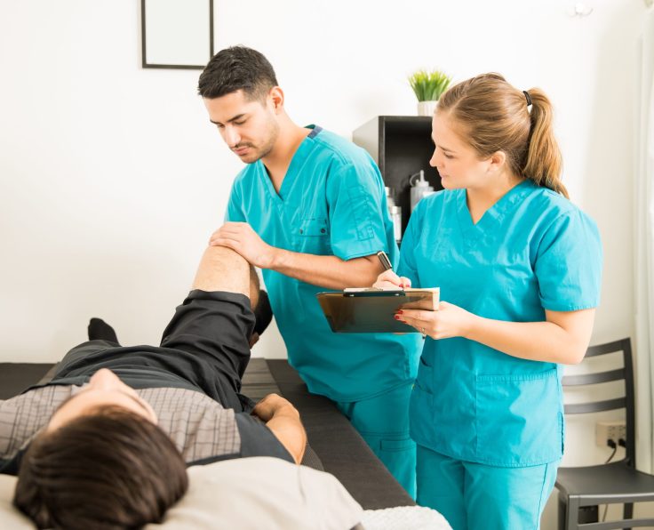 Young physiotherapist examining customer's leg while colleague noting in clipboard at hospital